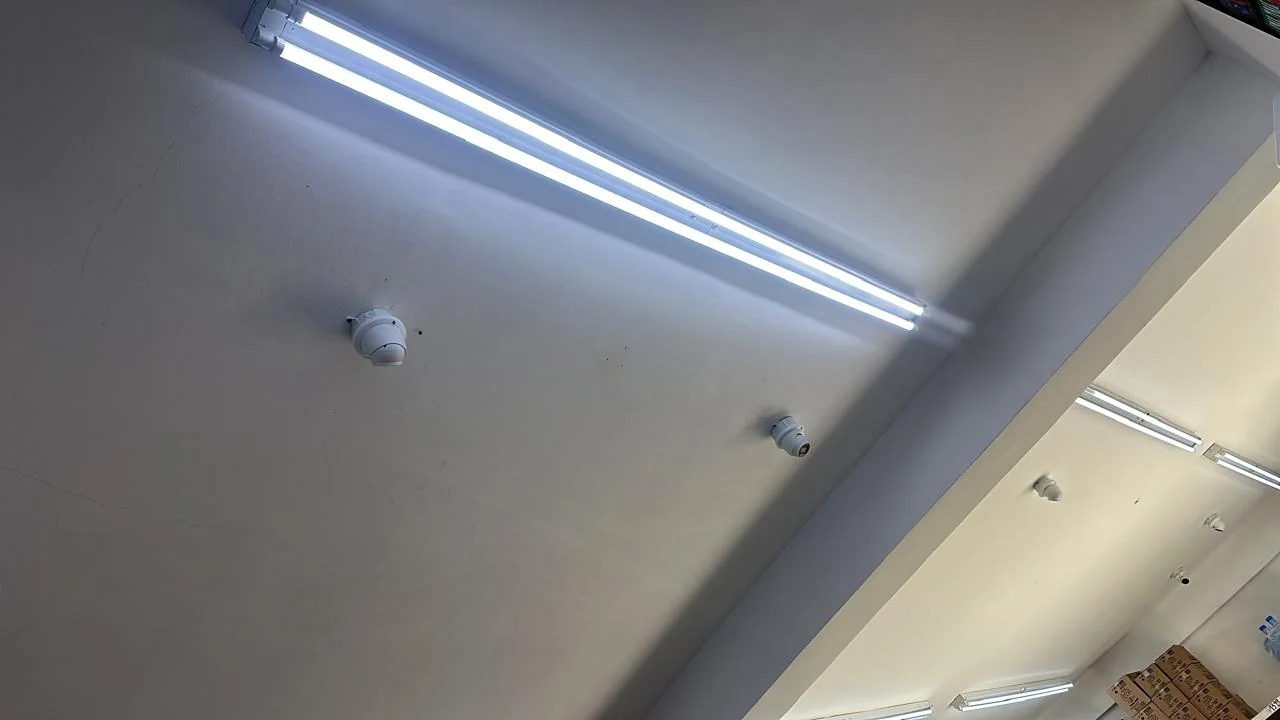 Two white surveillance cameras mounted on an interior ceiling beside a long fluorescent light.