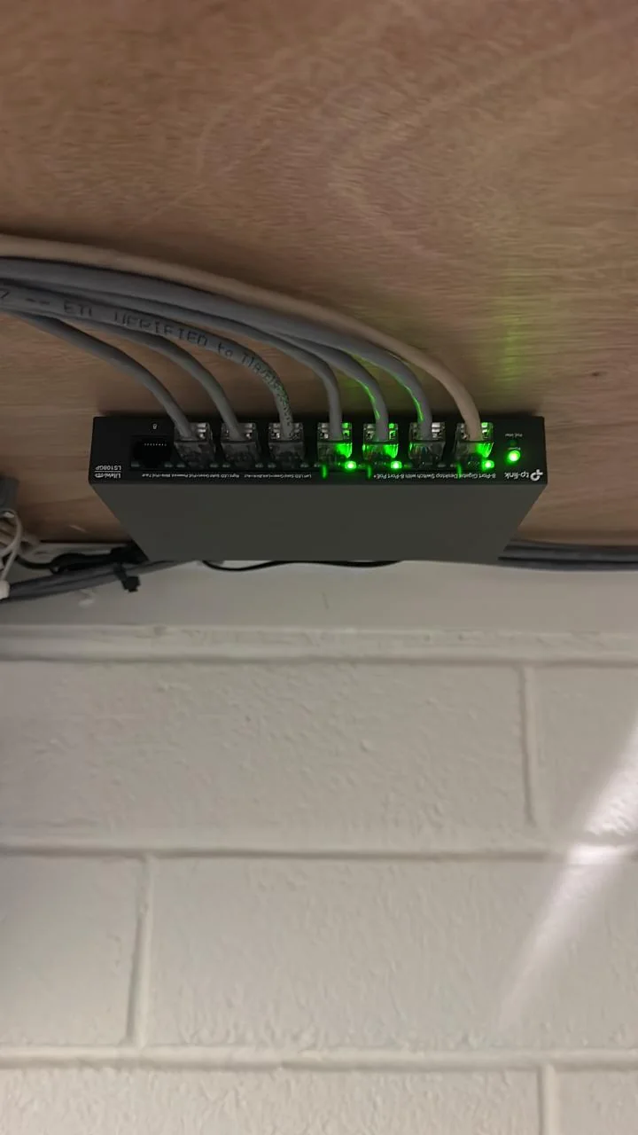 Compact network switch mounted below a wooden surface with connected cables and green indicator lights.