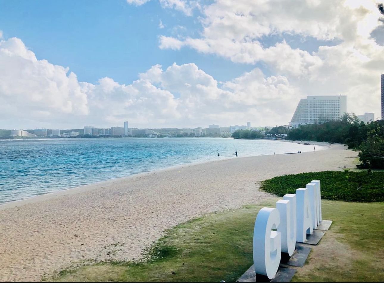 Client-supplied coastal photograph showing a sandy beach, blue water, distant buildings, and a Guam sign.