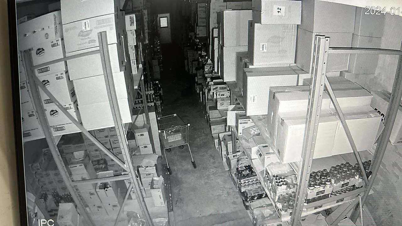 Monochrome surveillance view of a storage aisle with shelving and equipment.