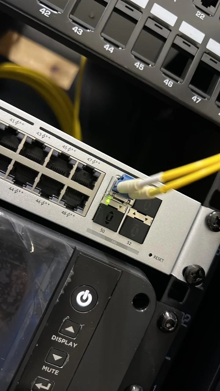 Network switch with Ethernet ports and a yellow fiber uplink cable connected beside rack equipment.