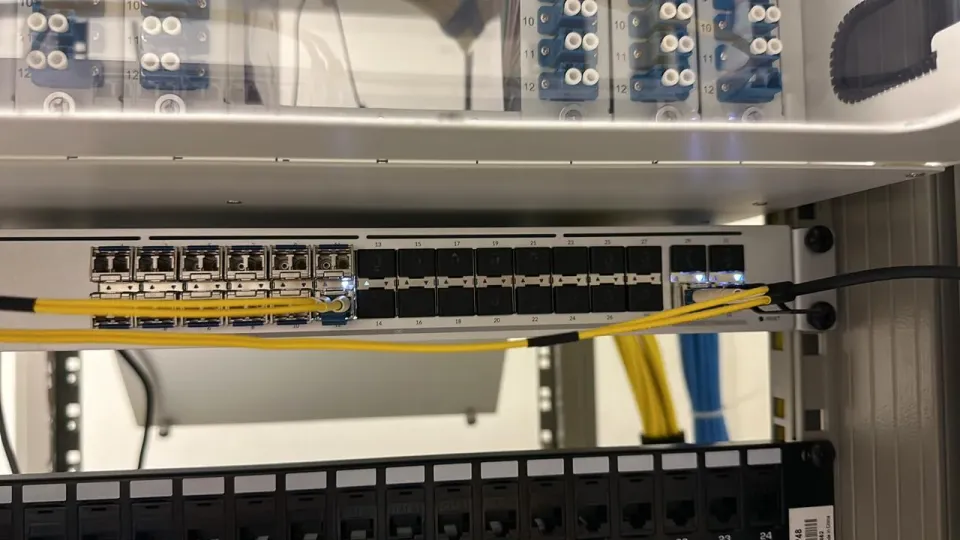 Rack-mounted optical switch with transceiver slots and yellow fiber patch cables below a fiber distribution panel.