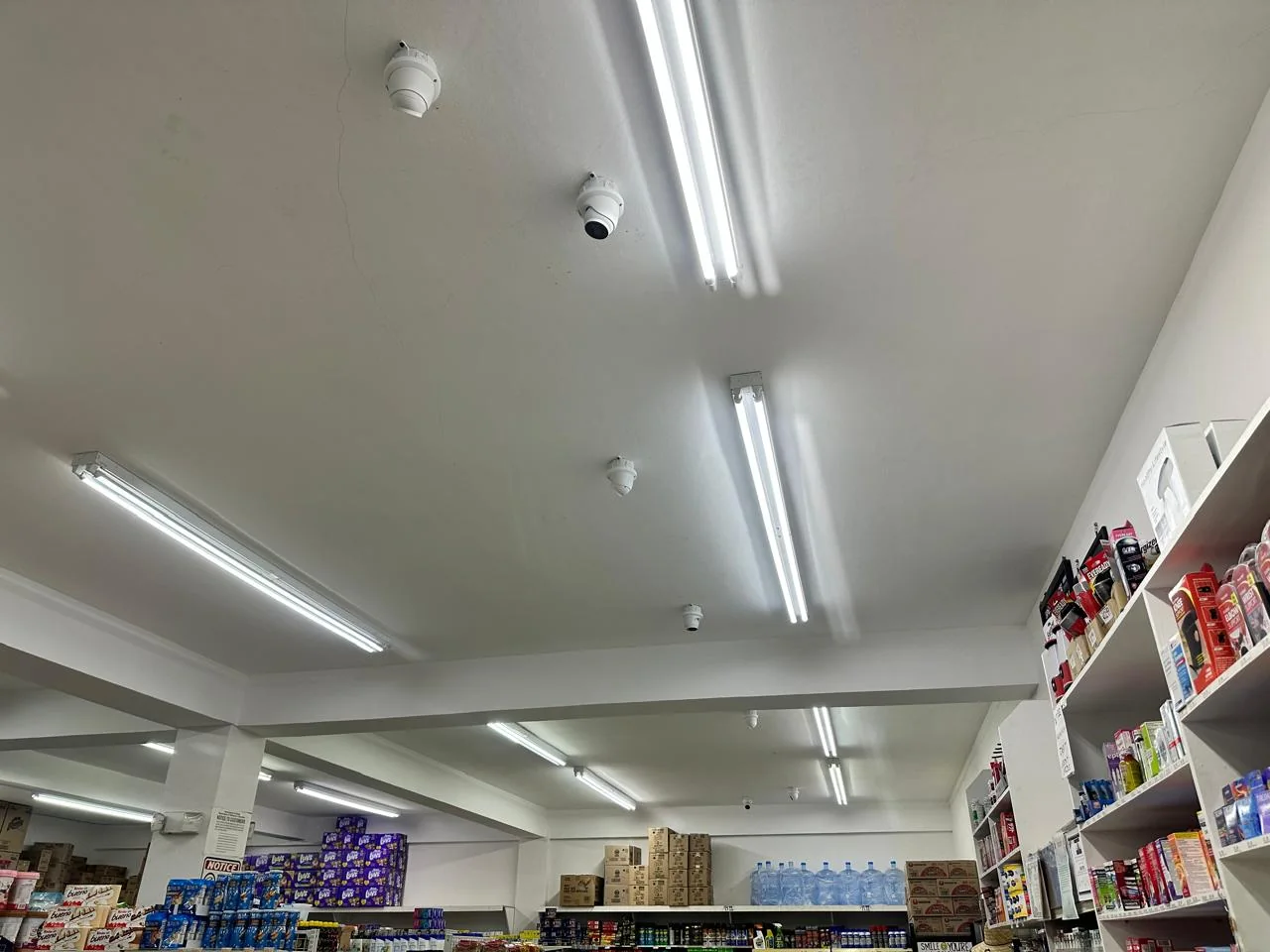 White surveillance camera mounted on a retail ceiling between fluorescent lights above store shelving.